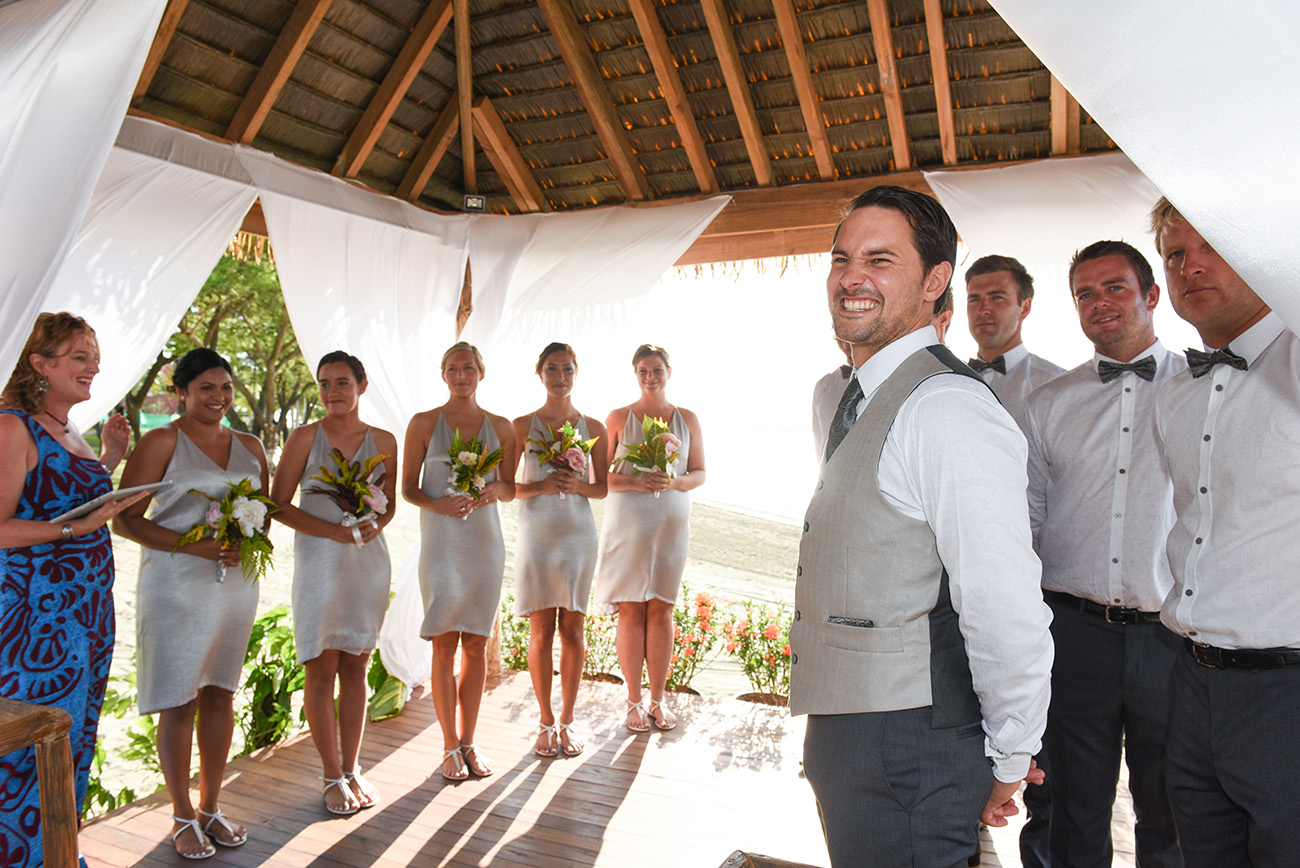 Under the beach hut the groom smiling when seeing arrival of his future wife surrounded his groomsmen and bridesmaids