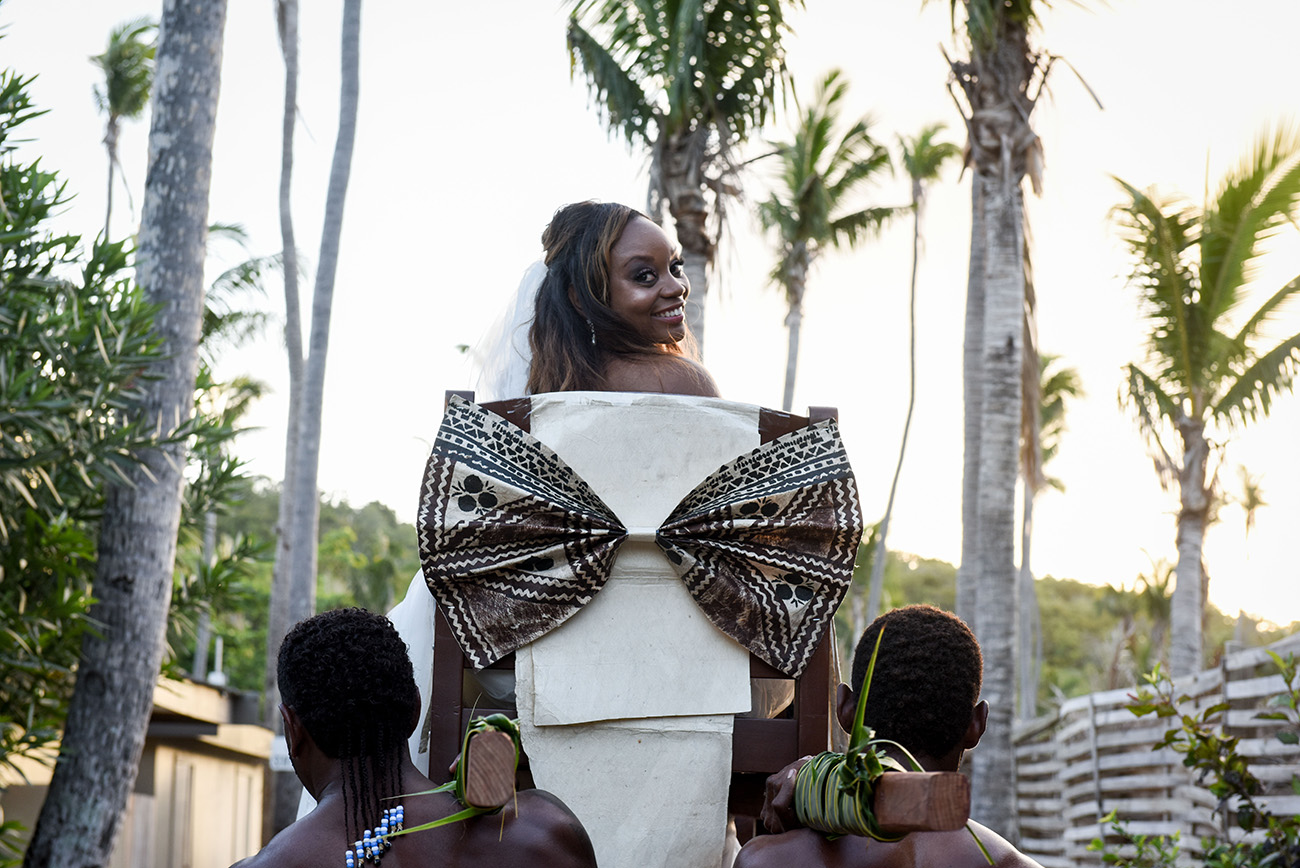 The bride is looking back at the photographer while sited on her chair carried by Fijian warriors at Paradise Cove Island Resort in the Yasawas, Fiji by Anais Photography