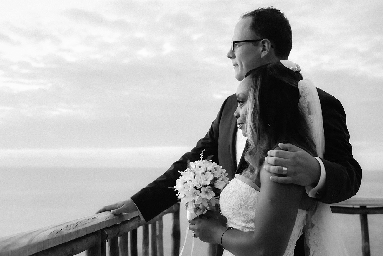 The groom is having his arm around his wife's while they are watching at the horizon at Paradise Cove island resort in the Yasawas in Fiji photographed by Anais Photography Black and white photograph
