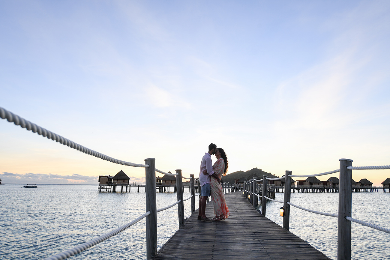 Newly wed couple kiss against villas on wooden bridge at sunset