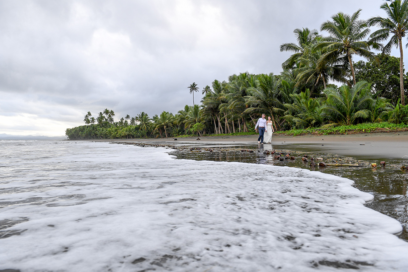 Stunning wideshot of bride and groom standing on the edge of sea and palm trees