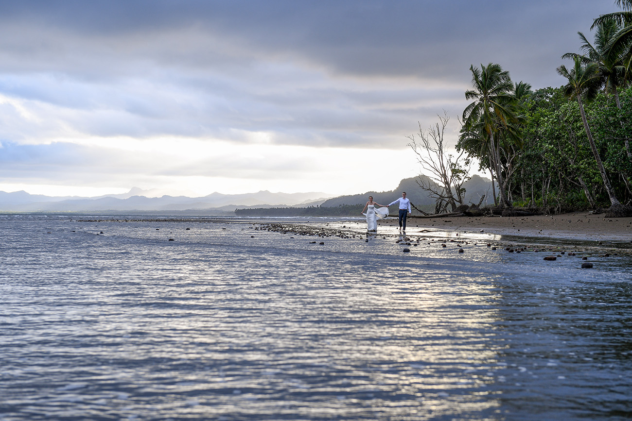 Bride and groom walk between palm trees and the Pacific ocean
