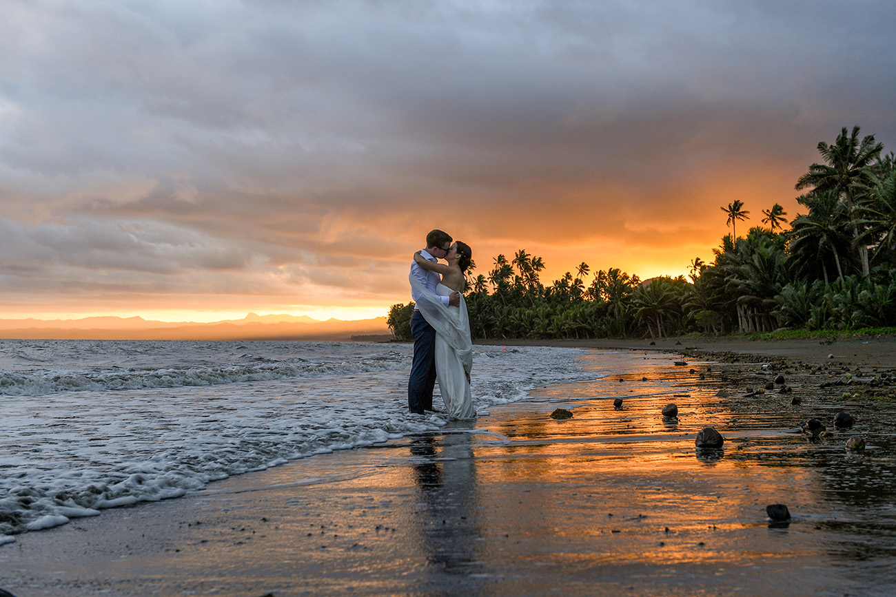 Wideshot of bride and groom at the sea kissing against the Fiji sunset