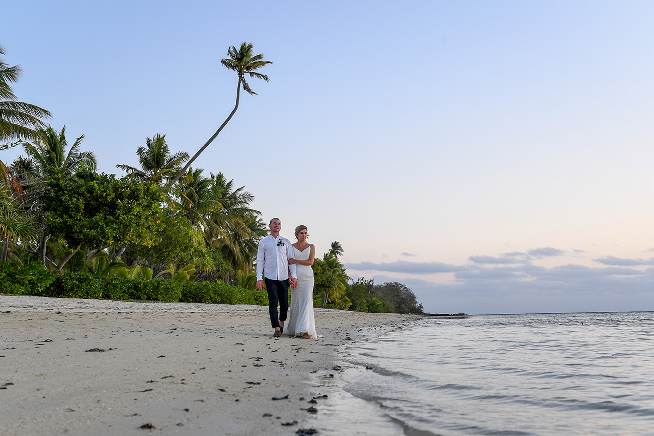 Bride and groom stroll along the sea against overhanging palm trees.