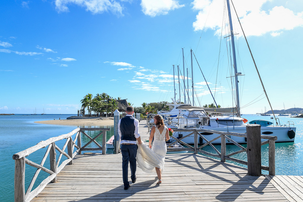 The bride holds the bride's train as they walk on the dock at Musket cove
