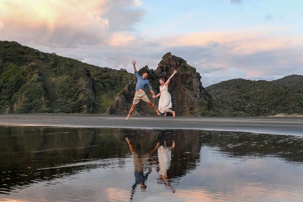 The newly weds leap in the air over the shallow water at Karekare
