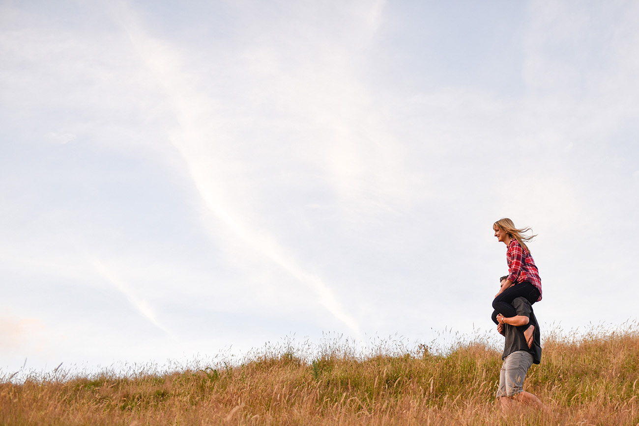 Profile piggy-back ride of the engaged couple in the fields Summer Country Engagement photo shoot Pukekokhe Auckland Photographer Anais Chaine