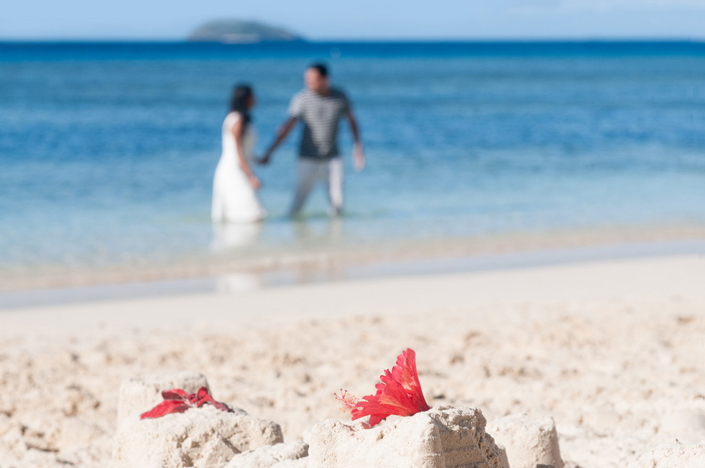 Blurred couple in the background holding hand in the sea water and walking to the beach with send castle in the front at Mana Island resort, Fiji by Anais Photography
