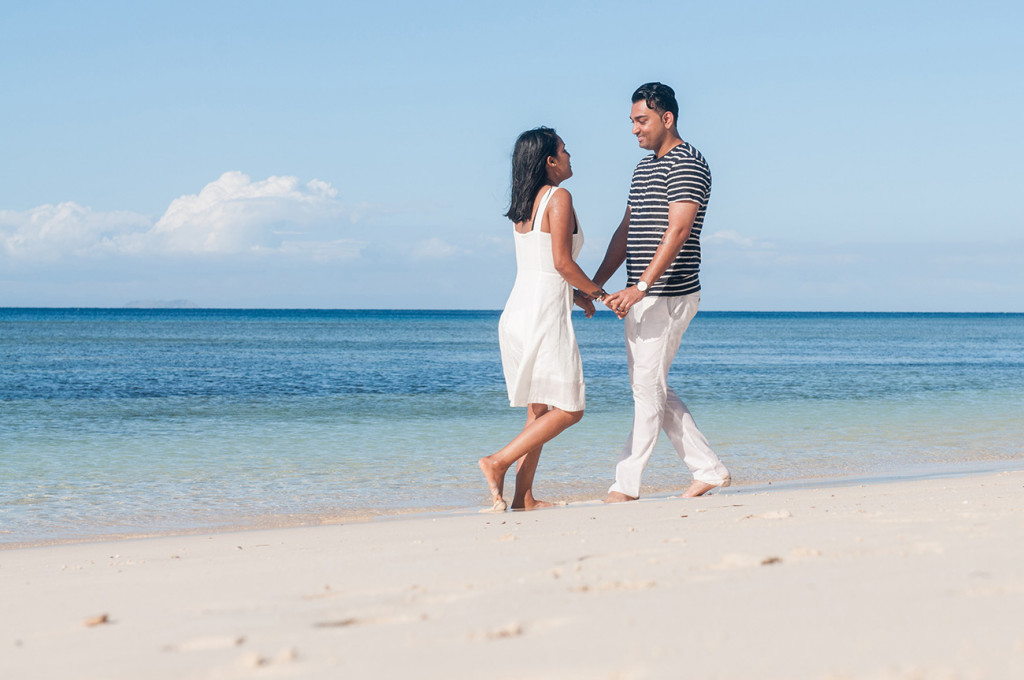 Couple by the beach with women wearing a white dress and the man a white trouser and a stripe blue shirt at Mana Island resort, Fiji by Anais Photography