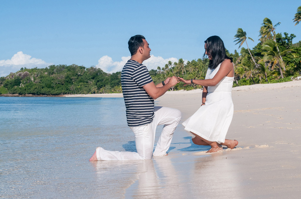 The man is putting the ring on the finger of his fiancee by the beach of Mana Island resort, Fiji by Anais Photography