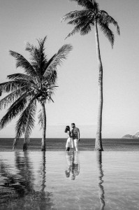 Black and white photograph with a couple between two palm trees by the swimming-pool and the ocean on the background at Mana Island resort, Fiji by Anais Photography