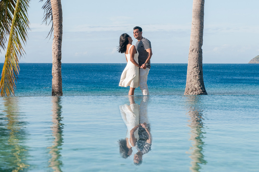 Couple looking at each others holding hands between two palm trees with their reflection in the pool and the ocean in the background at Mana Island resort, Fiji by Anais Photography