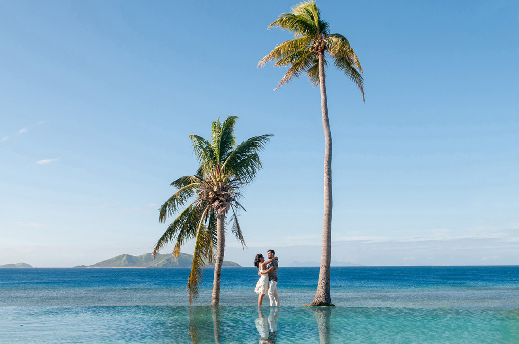 Couple cuddling by the pool between palm trees with ocean in the background at Mana Island resort, Fiji by Anais Photography
