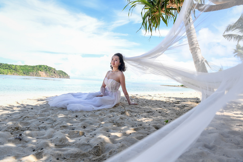 Bride sitting at the beach with her veil coming to the lens Paradise Cove island resort, Fiji