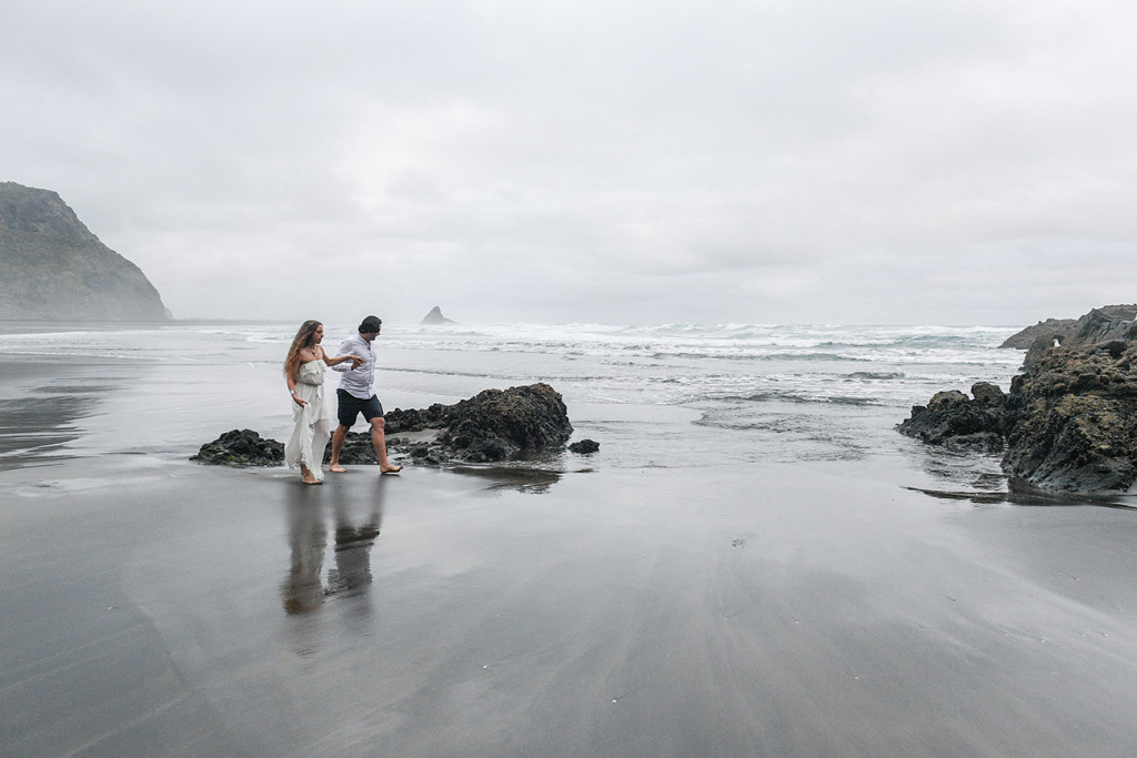 Eloped couple stroll against dull weather on black sand beach Karekare NZ Auckland