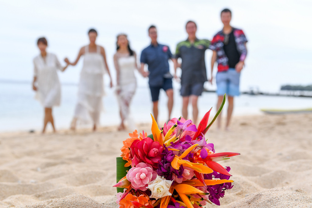 Married couple and wedding guests walk on beach with orange and pink bouquet sharp in the foreground