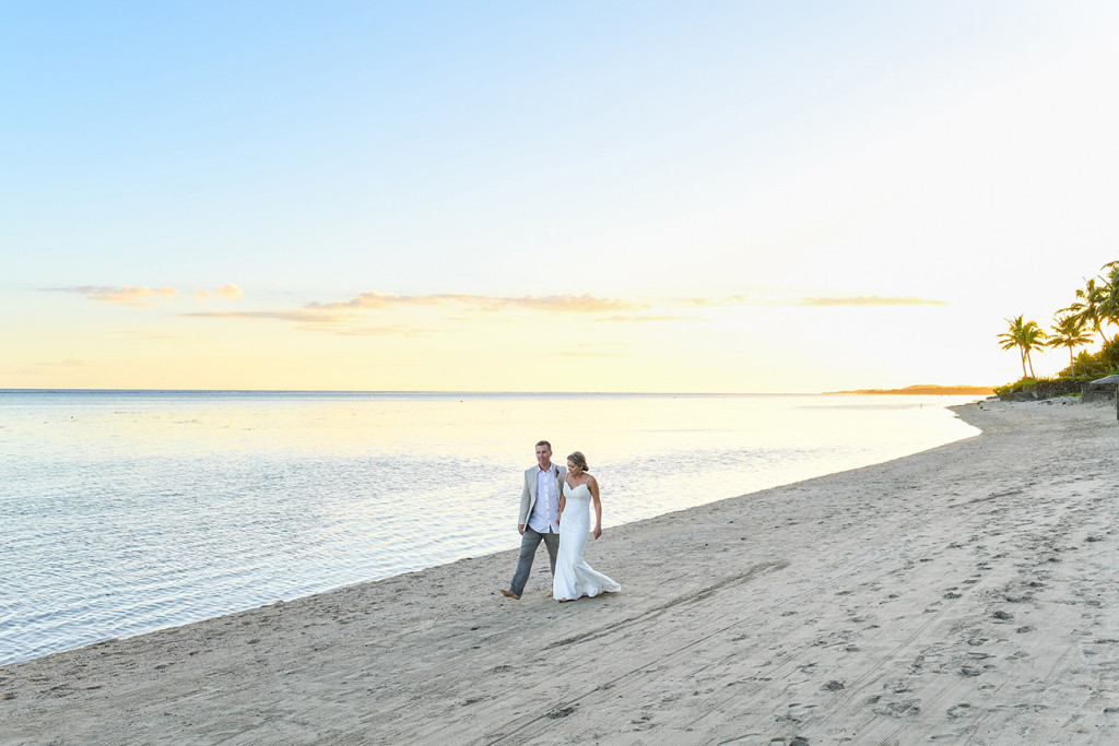 Breathtaking wide shot of bride and groom walking on beach against golden sunset