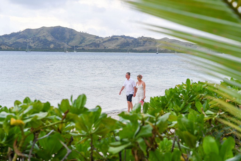 Long shot through palm trees of Bride and groom strolling against Fiji sea