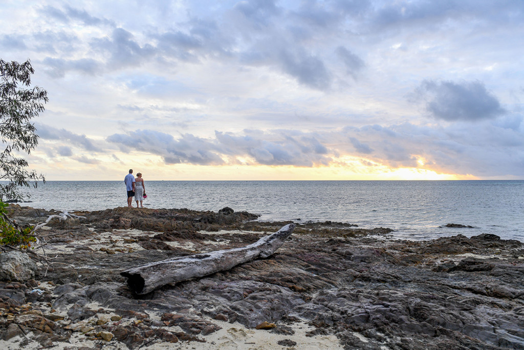 Wide landscape shot of Bride and groom standing on coral rocks against fiery sunset