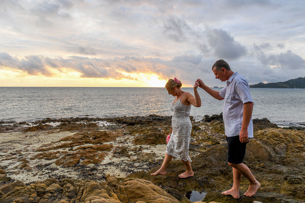 Groom helps bride walk on rocks by the sea