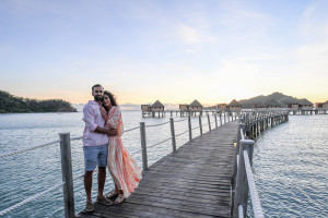 Newly married couple hug while on wooden bridge in front of Likuliku villas and Sunset