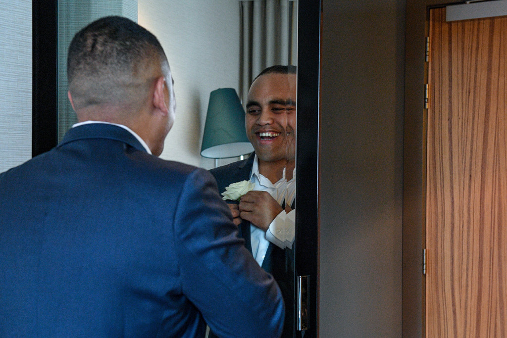 Groom smiles at mirror while adjusting his boutonniere