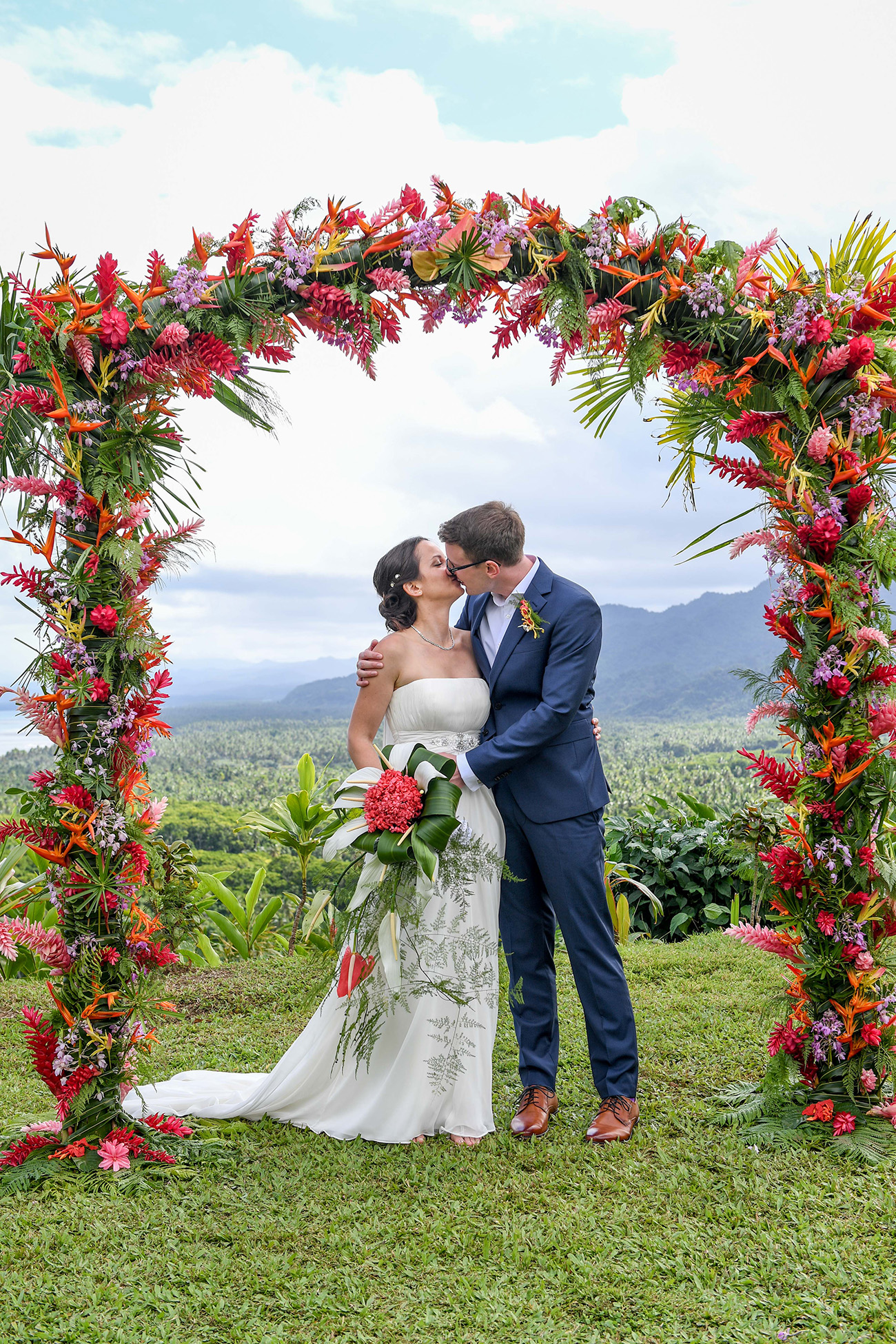Bride and groom kiss at the altar beneath the floral arc of red and pink flowers at the altar
