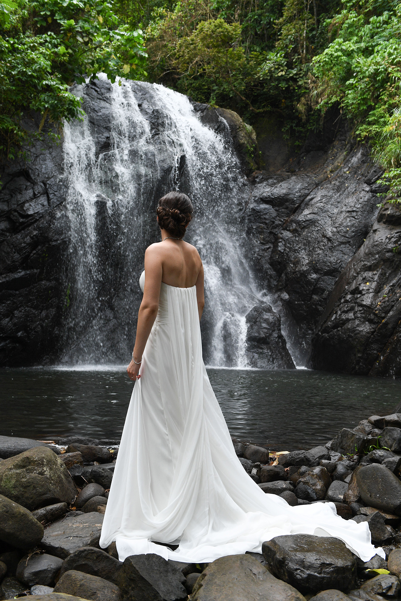 The bride in long white wedding dress stands next to waterfall