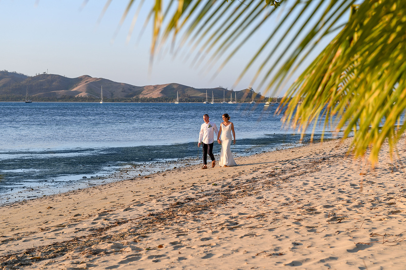 Bride and groom stroll hand in hand by the beach amongst palm trees