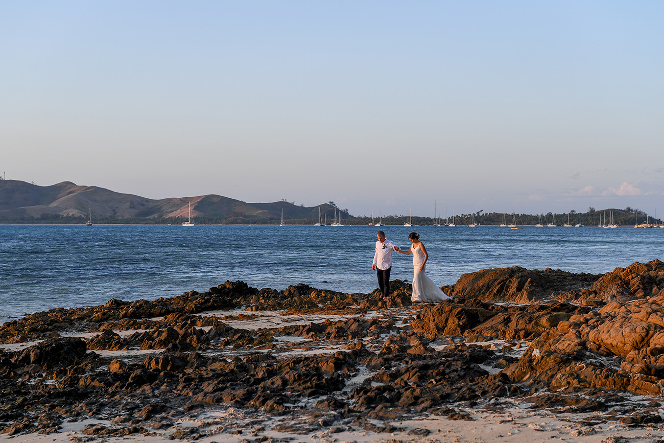 Wideshot of Bride and groom walking on coral rock against the sea