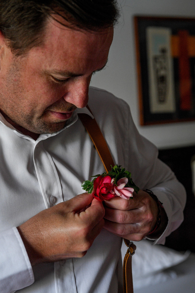 Groom adjusts pink tropical flower boutonniere on his suspenders