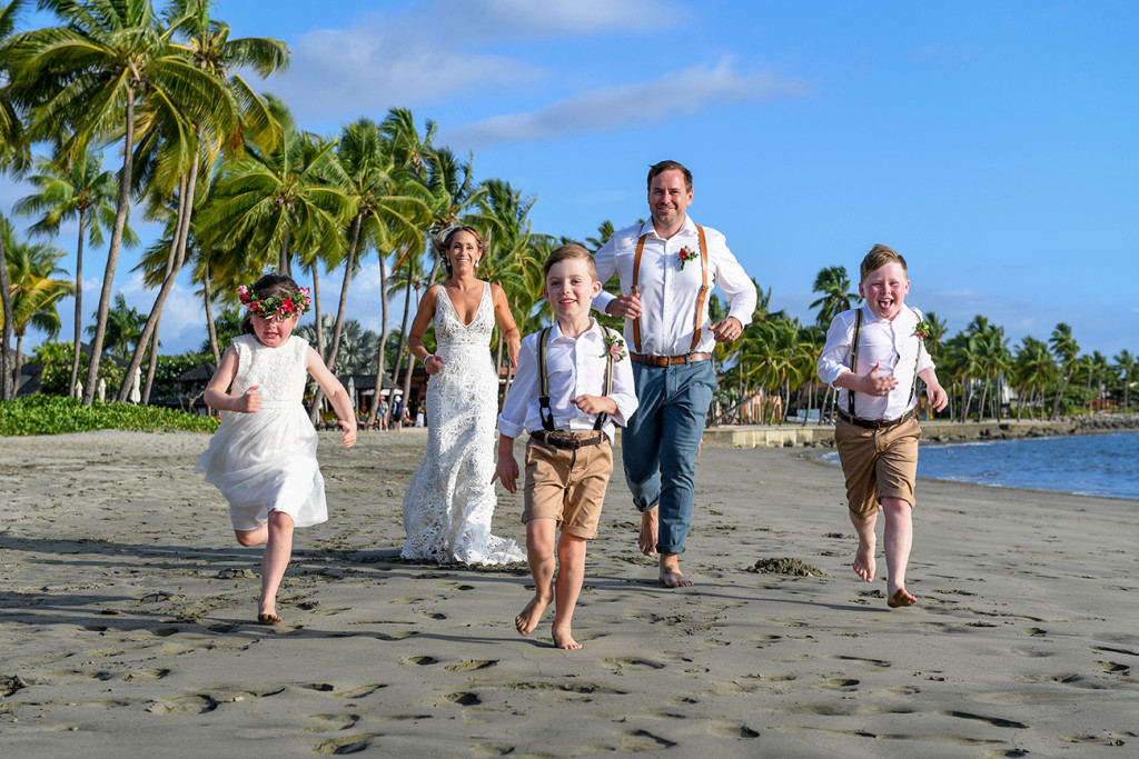 Newly married couple run on black sand beach with their family