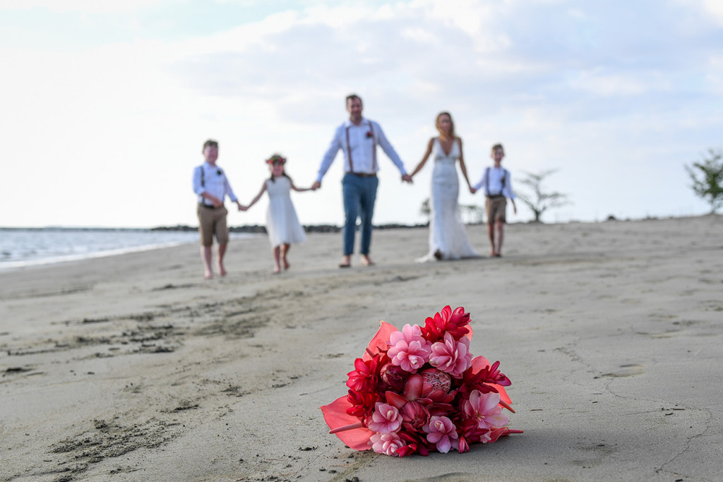 Bride, groom and family hold hands on the beach, tropical floral bouquet sharp in foreground