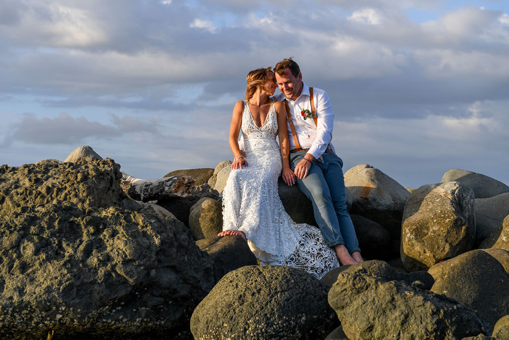The bride and groom have a moment seated on rocks in the sunset glow of the Pacific ocean