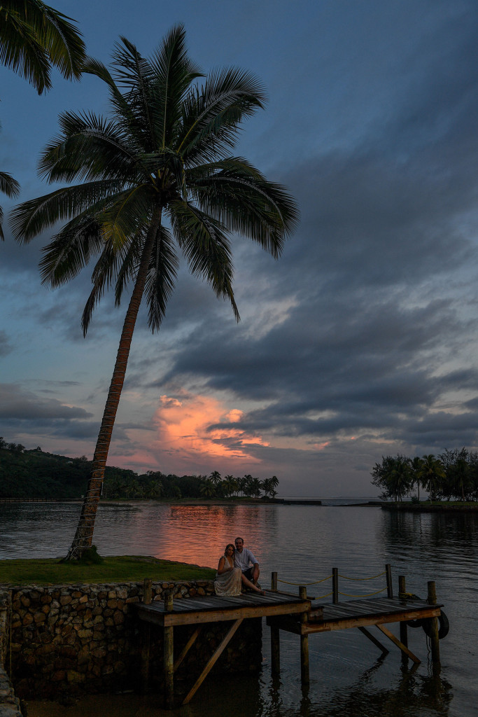 Bride and groom seated on bridge against Lone palm tree in fiery sunset