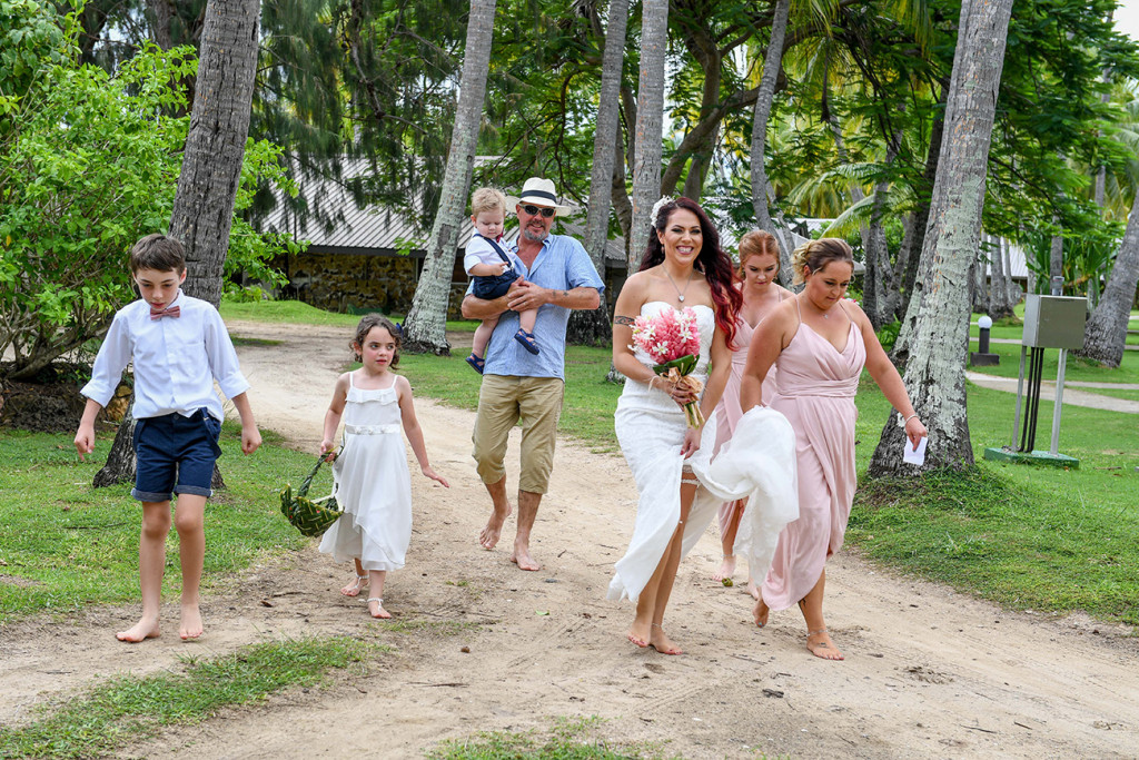 The bride, bridesmaids and flower girls and boys walk in the sand toward the ceremony.