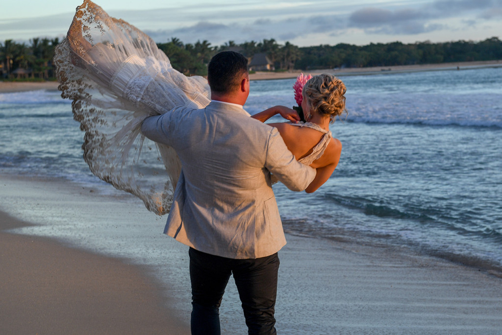 Groom lifts his wife in the air while strolling on the beach