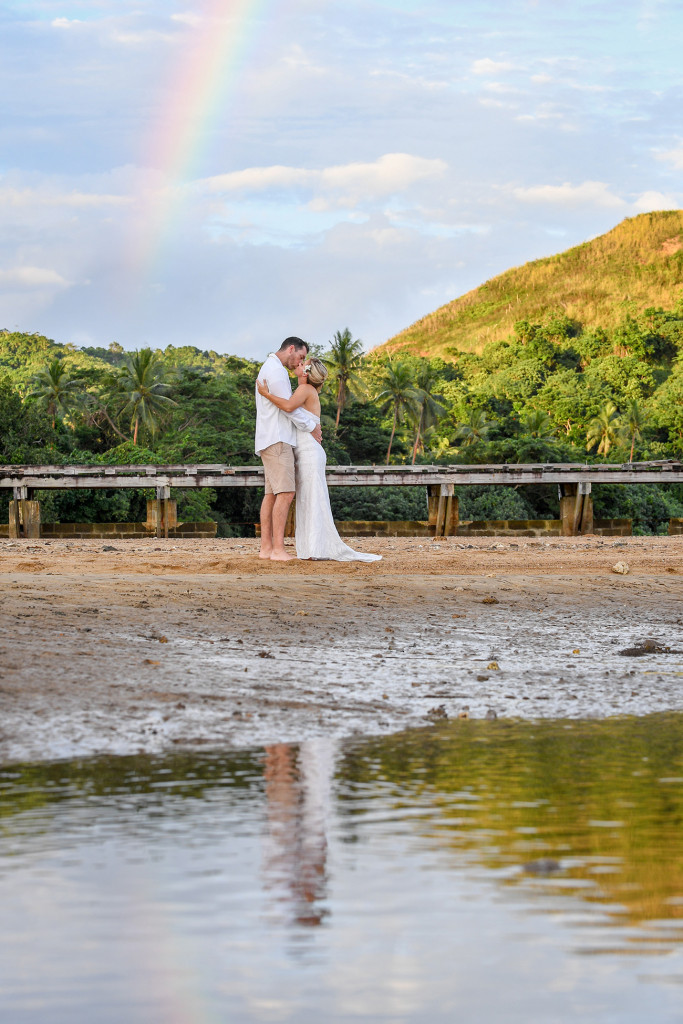 The bride and groom kiss under a rainbow at sunset