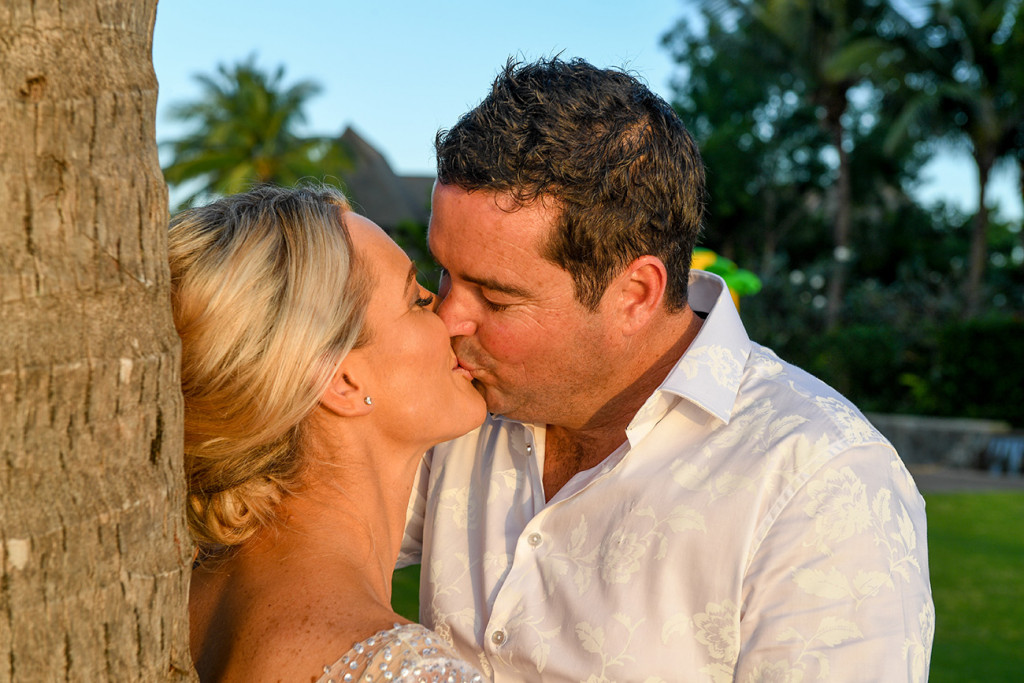 The sun glows on the couple as they kiss against a palm tree