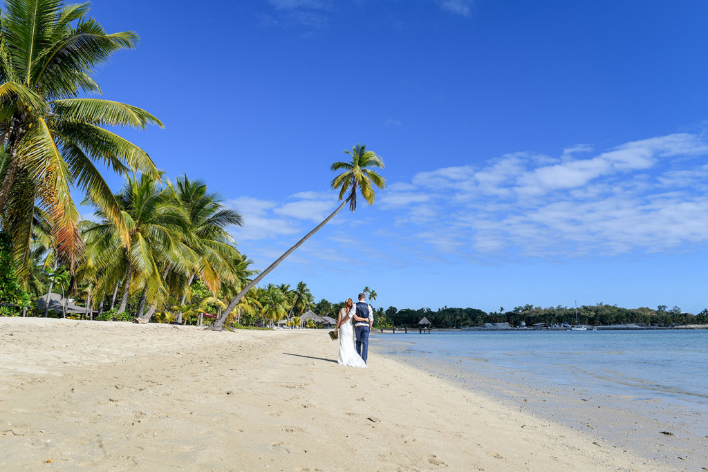 Palm trees sway over the married couple as they walk hand in hand on the Musket Cove beach