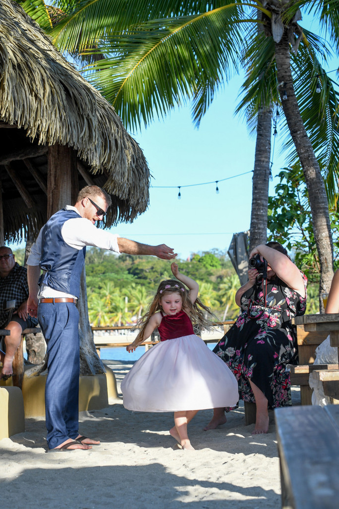 The flower girl twirls as she dances with the groom under the palm trees