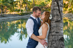 Reflections bounce off the sea as the groom cuddles his bride against stocky palm tree