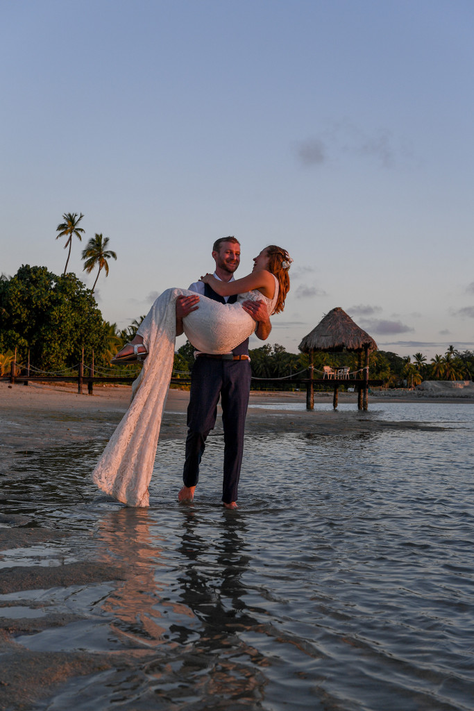 The groom whisks his bride away into the ocean at sunset