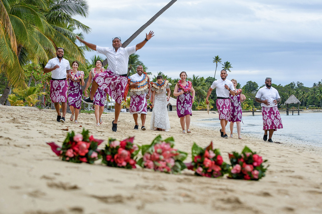 A groomsman leaps into the air while running towards the camera on the beach