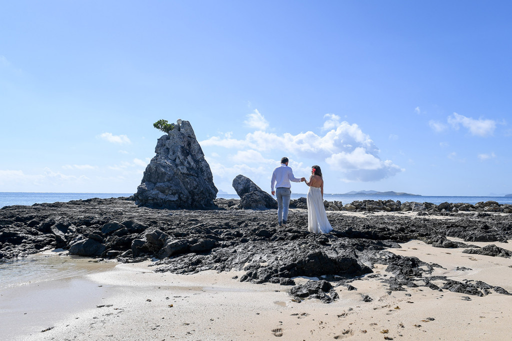 The couple holds hands while standing on the rocks at the Castaway reef against baby blue skies