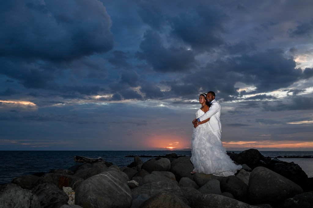 The bride and groom stand on the rocky dock at a pink sunset