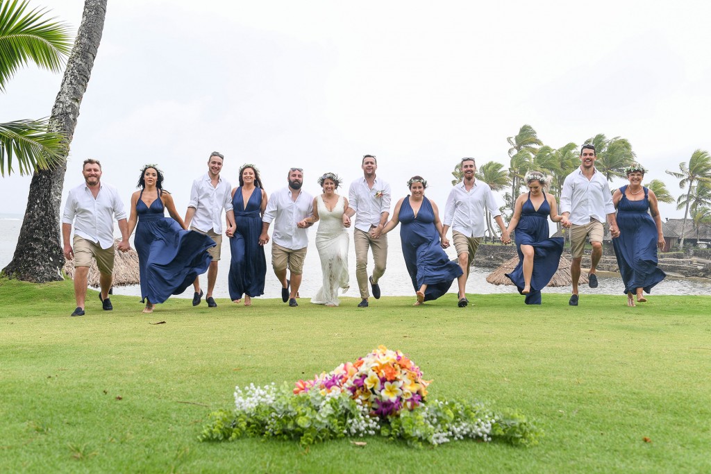 The bridal party run towards the camera with a bouquet in the foreground