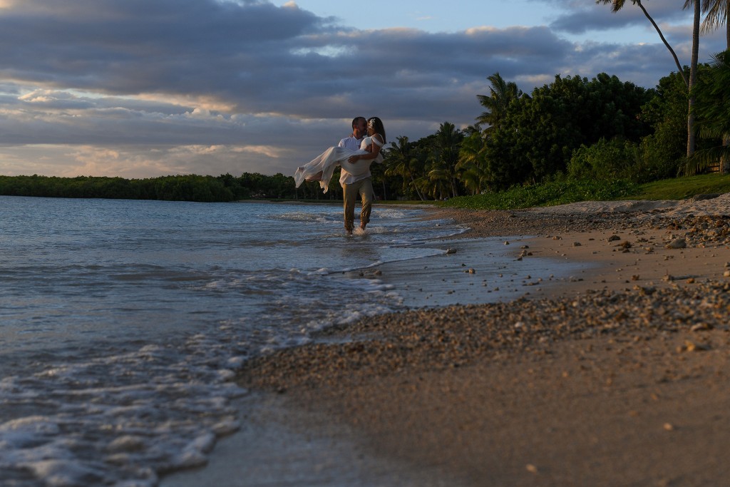 The groom carries his bride as he wades in the ocean at sunset
