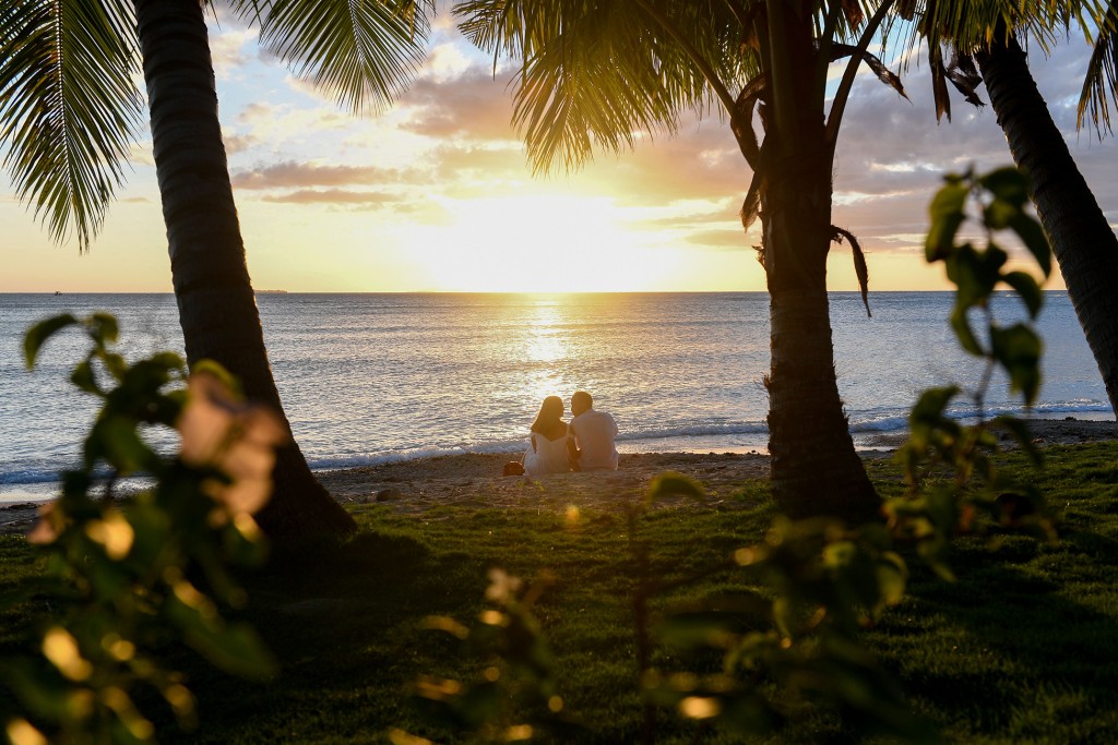 The loving couple sit between palm trees and stare into the golden Fiji sunset
