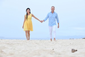 The engaged couple walk hand-in-hand on the beige beach against baby blue skies